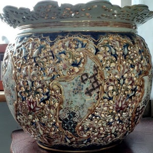 Puede incluir: Un macetero grande de cerámica blanca con intrincados diseños florales dorados y azules. El macetero tiene un diseño delicado y calado y un borde festoneado.