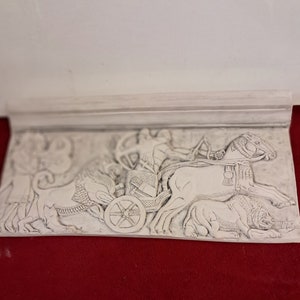 May include: A white plaster relief sculpture depicting a chariot pulled by two horses. The chariot is driven by a figure holding reins. The sculpture is detailed with intricate carvings and is mounted on a white plaster base.