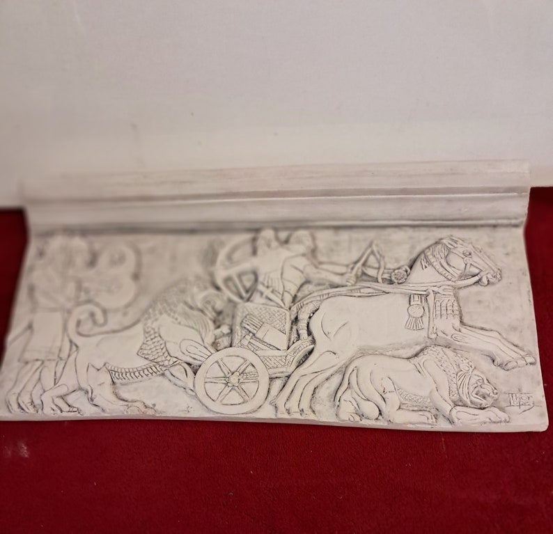 May include: A white plaster relief sculpture depicting a chariot pulled by two horses. The chariot is driven by a figure holding reins. The sculpture is detailed with intricate carvings and is mounted on a white plaster base.