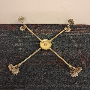 May include: A gold-toned metal table base with an X-shaped design. Each end features a decorative floral element. The center has a circular plate. The base is set against a dark, textured background.