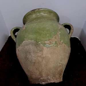 May include: A large, antique green and tan ceramic vase with two handles. The vase has a wide body and a narrow neck. The green paint is peeling in areas, revealing the tan ceramic underneath. The vase is likely an antique decorative item.