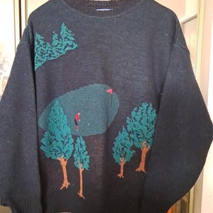 May include: Dark navy blue sweater with a golf course design. The sweater features a green golf course with a red flag, a golfer, and green trees with brown trunks. The sweater is hanging on a wooden hanger.