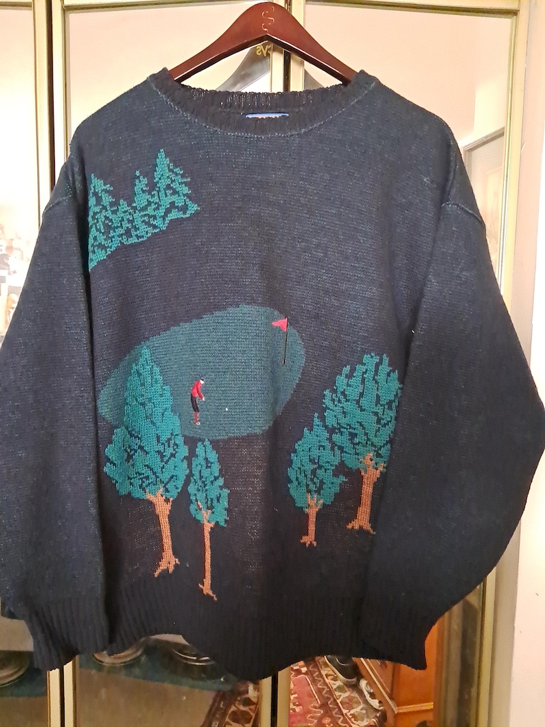 May include: Dark navy blue sweater with a golf course design. The sweater features a green golf course with a red flag, a golfer, and green trees with brown trunks. The sweater is hanging on a wooden hanger.