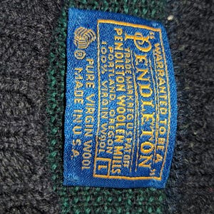 May include: Close-up of a blue label with gold text from Pendleton Woolen Mills. The label reads "WARRANTED TO BE A" and "PURE VIRGIN WOOL". The label is sewn onto a dark blue and green wool fabric.