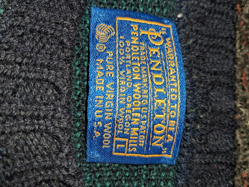 May include: Close-up of a blue label with gold text from Pendleton Woolen Mills. The label reads "WARRANTED TO BE A" and "PURE VIRGIN WOOL". The label is sewn onto a dark blue and green wool fabric.