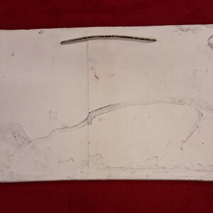 May include: A white, rectangular, ceramic tile with a crack running across the surface. The tile has a rough, textured surface and appears to be old and worn.