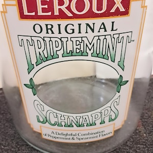 Könnte beinhalten: Eine klare Glasflasche mit einem weißen und roten Etikett. Auf dem Etikett steht "LEROUX ORIGINAL TRIPLEMINT SCHNAPPS" in grüner und weißer Schrift. Auf dem unteren Teil des Etiketts steht "A Delightful Combination of Peppermint & Spearmint Flavors."