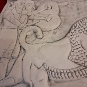 May include: A close-up of a white plaster relief sculpture depicting two figures with beards, one holding a staff, and a third figure in a cloak. The figures are standing on a stylized animal with scales.