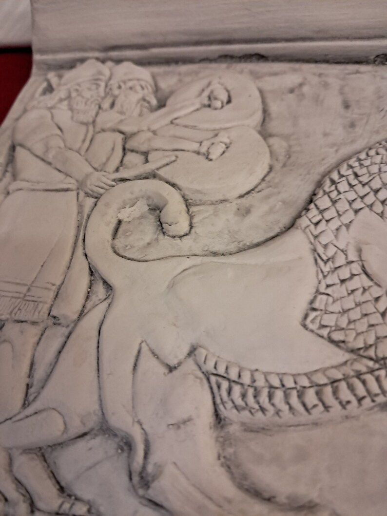 May include: A close-up of a white plaster relief sculpture depicting two figures with beards, one holding a staff, and a third figure in a cloak. The figures are standing on a stylized animal with scales.