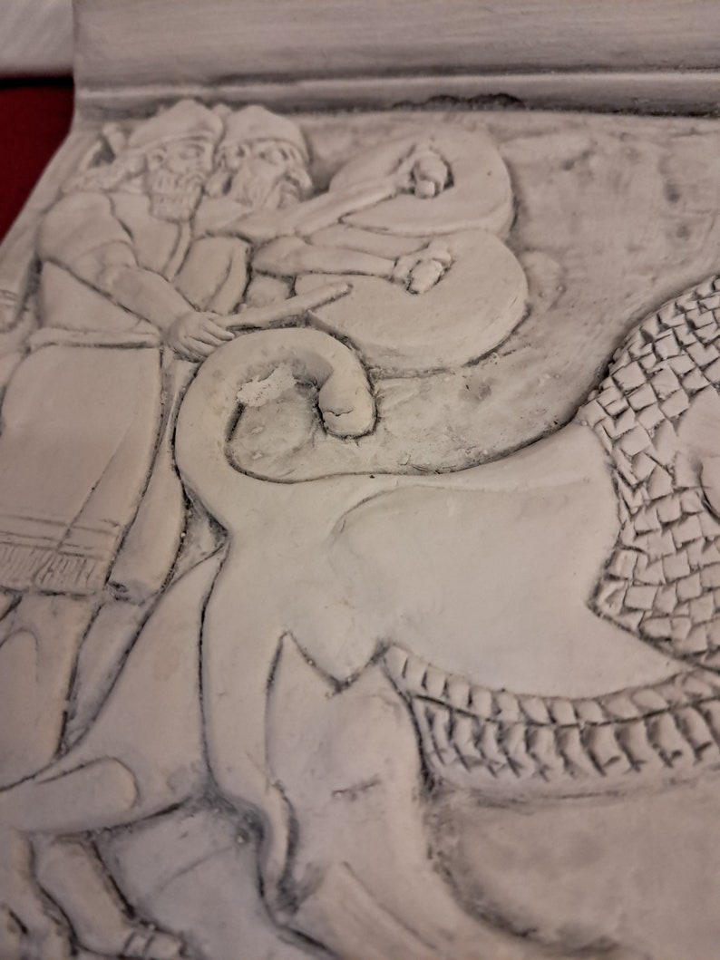 May include: A white plaster relief sculpture depicting three figures, possibly men, standing in front of a large, stylized animal. The figures are wearing long robes and have their arms raised. The animal has a scaly body and a large head with a wide mouth.