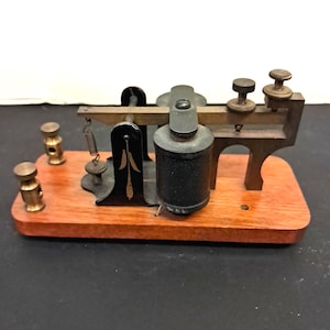 May include: A telegraph key mounted on a rectangular wooden base. The device features a black metal upright with a gold leaf design, a cylindrical black coil, a brass lever arm, and two brass binding posts with knurled adjustment screws.