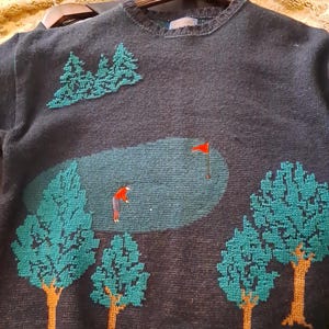 May include: Dark navy blue sweater with a golf course design. The sweater features a green golf course scene with a golfer, trees, and a red flag. The trees have teal green leaves and brown trunks.
