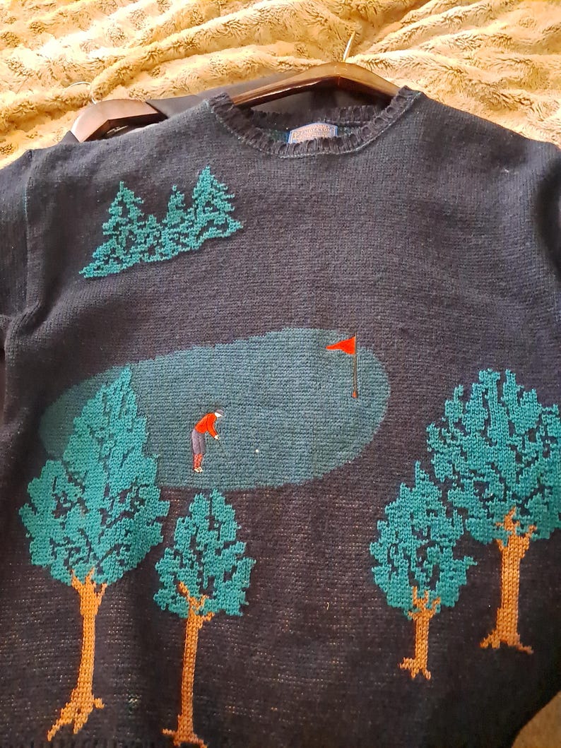 May include: Dark navy blue sweater with a golf course design. The sweater features a green golf course scene with a golfer, trees, and a red flag. The trees have teal green leaves and brown trunks.
