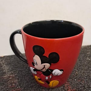 May include: A red ceramic mug featuring a classic Mickey Mouse design. The mug has a black handle and interior, with Mickey Mouse in his iconic pose. A fun and colorful item for Disney fans.