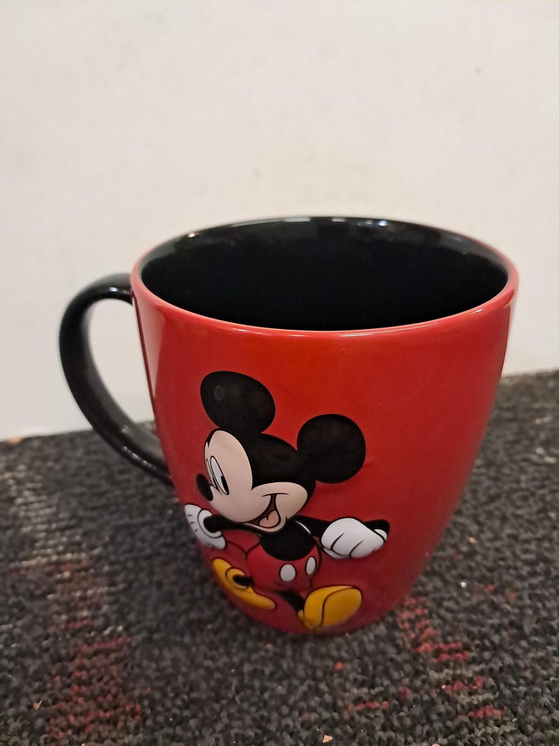May include: A red ceramic mug featuring a classic Mickey Mouse design. The mug has a black handle and interior, with Mickey Mouse in his iconic pose. A fun and colorful item for Disney fans.