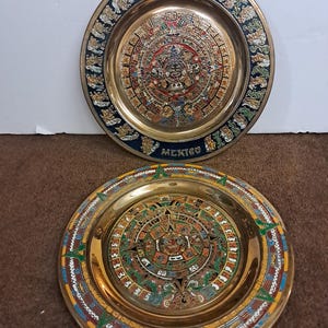 May include: Two decorative brass plates with intricate Aztec calendar designs. The plates feature colorful details and the word "MEXICO" at the bottom. The plates have a gold finish and are displayed on a brown surface.