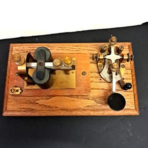 May include: A vintage telegraph key mounted on a rectangular wooden base. The device features brass components, two black cylindrical coils, and a black circular knob. The mechanism includes various metal screws, adjustment nuts, and a metal lever arm.