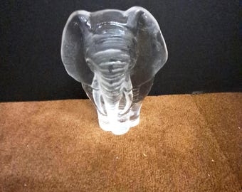 May include: A clear glass elephant figurine with detailed trunk and ears. The elephant is standing upright, showcasing its form. The figurine is on a brown surface with a black background.