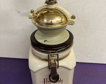 Puede incluir: Un molinillo de café de cerámica blanca con un mango de madera y un molinillo de metal dorado. El molinillo tiene una tapa marrón con un cierre de metal y dice "Harrods Knightsbridge Coffee" en la parte delantera.