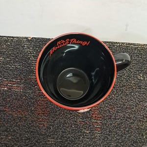 May include: Black ceramic mug with a red rim and a black handle. The interior is black, and the words "It's a Mouse Thing!" are written in red script around the rim. The mug is sitting on a dark gray surface.