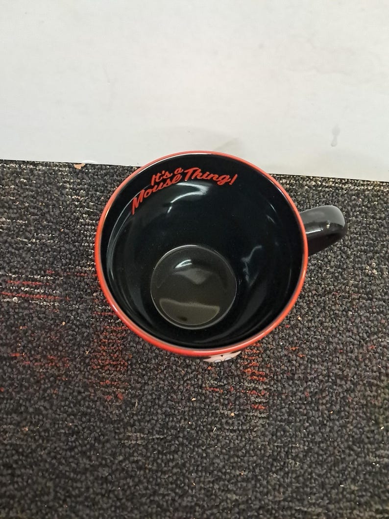 May include: Black ceramic mug with a red rim and a black handle. The interior is black, and the words "It's a Mouse Thing!" are written in red script around the rim. The mug is sitting on a dark gray surface.