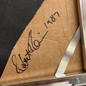 Op de afbeelding: Een close-up van de achterkant van een ingelijst kunstwerk, met een bruine papieren achterkant met een handgeschreven zwarte inkt handtekening en de datum 1987. De lijst is gemaakt van zilverkleurig metaal, met een zichtbare hoek en metalen clips.