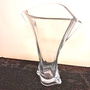 May include: A clear glass vase with a unique, flared design. The vase has a wide, open top and a sculpted base. The glass is transparent, allowing light to pass through. The vase is likely used for decorative purposes.