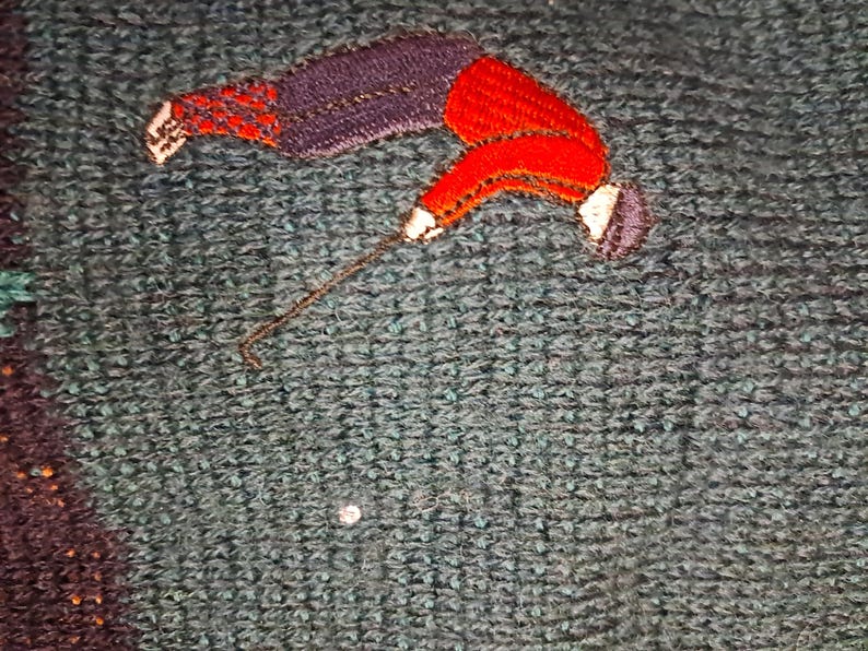 May include: Close-up of a textured, teal-colored fabric with an embroidered figure. The figure is depicted in a diving pose, with a red jacket, purple pants, and a white hat. The embroidery has gold thread accents.