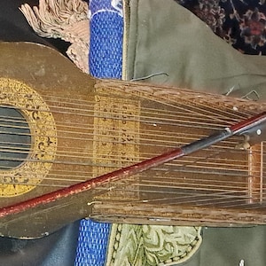 May include: An antique, stringed musical instrument with a dark, aged finish. The instrument features a round sound hole with decorative detailing and numerous strings. A bow rests across the strings, suggesting it is a bowed instrument. The instrument's design is ornate and vintage.