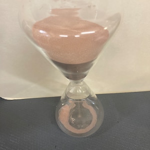 May include: A glass hourglass with brown sand. The sand is flowing from the top bulb to the bottom bulb.
