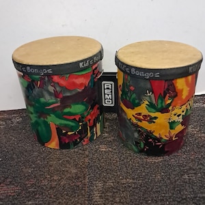 May include: A pair of Remo Kid's Bongos featuring tan drumheads and a colorful jungle-themed wrap with illustrations of animals and plants. The black rim of each drum is printed with the text Kid's Bongos, and a black Remo logo tag is attached.