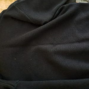 May include: A black sweater with a ribbed cuff and a tag. The sweater is made of a soft, knitted material. The image is taken from a slightly elevated angle, showing the texture and drape of the fabric.