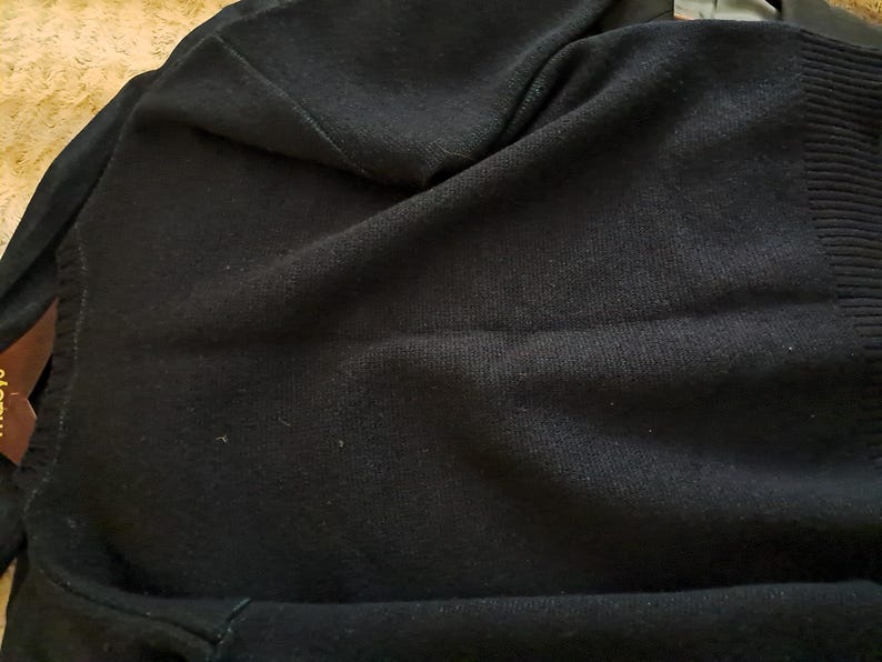 May include: A black sweater with a ribbed cuff and a tag. The sweater is made of a soft, knitted material. The image is taken from a slightly elevated angle, showing the texture and drape of the fabric.