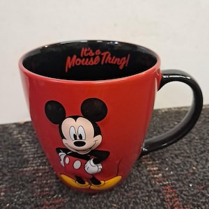 May include: Red ceramic mug featuring Mickey Mouse with a black handle and interior. The mug has the phrase "It's a Mouse Thing!" printed inside. The mug is a Disney-themed product.