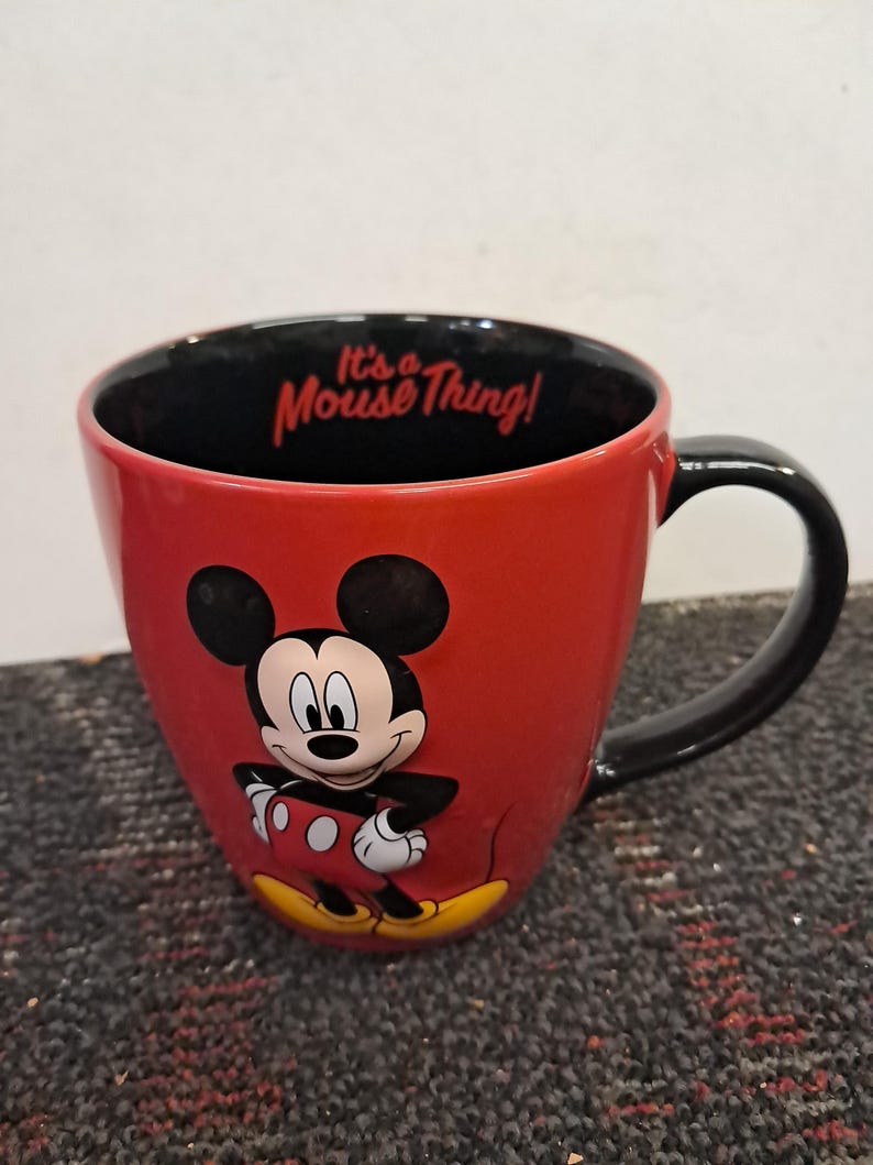 May include: Red ceramic mug featuring Mickey Mouse with a black handle and interior. The mug has the phrase "It's a Mouse Thing!" printed inside. The mug is a Disney-themed product.