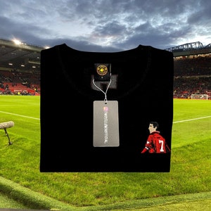 Puede incluir: Camiseta negra con una imagen bordada en rojo y blanco de un jugador de fútbol con el número 7. La camiseta tiene una pequeña etiqueta dorada con el texto "TWOYELLOWSOFFICIAL" en ella.