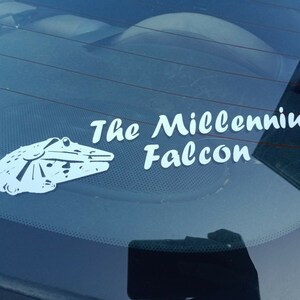 May include: A white vinyl sticker on a blue car window. The sticker features a silhouette of the Millennium Falcon spaceship from Star Wars and the text "The Millennium Falcon".