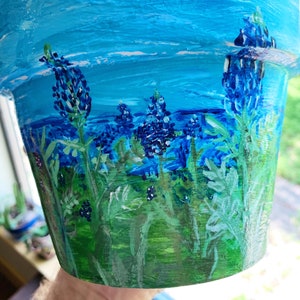 May include: A blue painted terracotta flower pot with a landscape scene of bluebonnets and green grass. The sky is painted in shades of blue with white clouds.