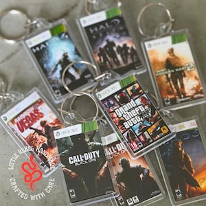 Peut inclure: Une collection de porte-clés rectangulaires présentant des couvertures miniatures imprimées de divers jeux vidéo Xbox 360, incluant des titres comme Grand Theft Auto V, Call of Duty Black Ops et Halo, chacun fixé à un anneau métallique circulaire.