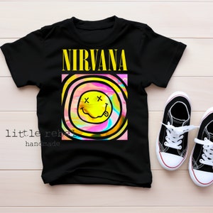 May include: Black t-shirt featuring a yellow smiley face graphic and the word "NIRVANA" in yellow lettering. The smiley face is set against a pink, blue and green background with black outlines.
