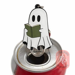 Peut inclure: Un accessoire de canette en forme de fantôme blanc lisant un livre vert, attaché à un anneau noir. Le fantôme a des yeux ovales noirs et est drapé dans un drap blanc. La canette est rouge et argentée.