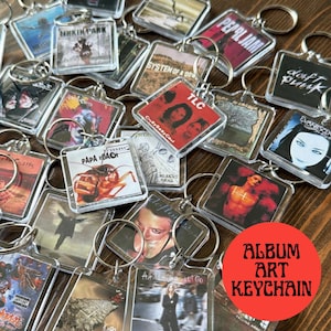 May include: A selection of clear acrylic keychains with album art from various bands, including Pearl Jam, Daft Punk, and Papa Roach. Each keychain has a silver metal ring. A red circle with the text "ALBUM ART KEYCHAIN" is visible in the image.