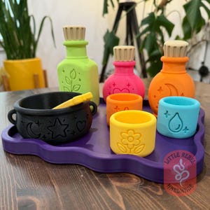 Peut inclure: Un ensemble de flacons de potion colorés, de petites tasses et un chaudron noir avec des gravures célestes et florales, disposés sur un plateau violet en forme de nuage. Les objets présentent des motifs en relief incluant lune, étoile, cœur, fleur et goutte.