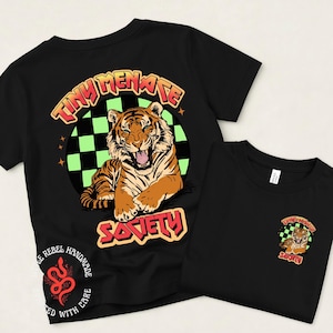 May include: A black t-shirt featuring a large graphic on the back and a smaller version on the front. The graphic shows a tiger in front of a green and black checkered circle with the text "TINY MENACE SOCIETY" in orange and red letters.
