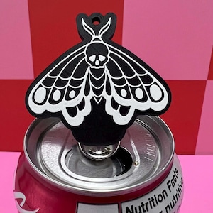 May include: A black and white moth-shaped can tab opener. The opener has a skull design on the moth's head and is placed on a red can. The background is a checkered pattern of pink and red.