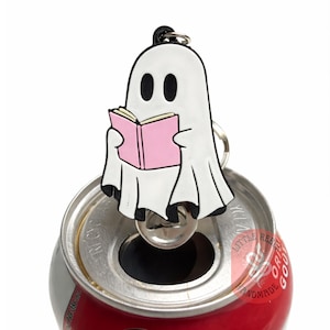 Peut inclure: Un porte-clés en forme de fantôme blanc lisant un livre rose, attaché à une canette de soda rouge. Le fantôme a des yeux noirs et un contour noir. La canette est ouverte, avec le porte-clés fantôme posé sur l'ouverture.