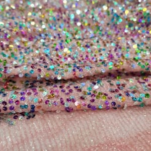 May include: Close-up of pink fabric covered in iridescent sequins. The sequins are a mix of colors, including pink, blue, green, and purple. The fabric has a textured appearance, with a soft, slightly fuzzy surface.