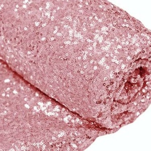 Könnte beinhalten: Eine Rolle aus rosa Paillettenstoff. Der Stoff hat eine hellrosa Farbe und ist mit kleinen, glänzenden Pailletten bedeckt. Der Stoff ist aufgerollt und zeigt die Textur und den Glanz des Materials. Ideal für Eventdekoration oder Bastelarbeiten.