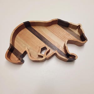 May include: A bear-shaped wooden tray constructed from alternating strips of light tan and dark brown wood. The tray features a hollowed-out center with raised edges, showcasing the natural grain patterns and color variations of the wood pieces.