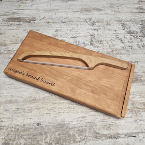 May include: A rectangular light brown wooden bread board with the engraved text "Angie's bread board" on the left side. A matching wooden bread knife with a thin metal wire blade is positioned diagonally across the surface of the board.